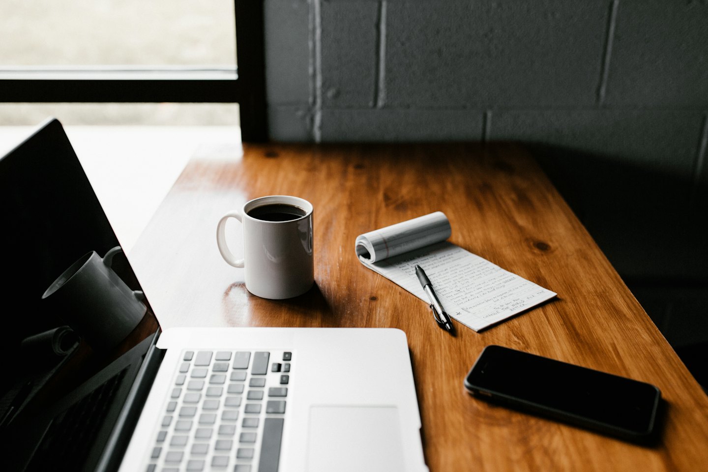 Laptop, coffee mug, and notepad on a wooden desk.